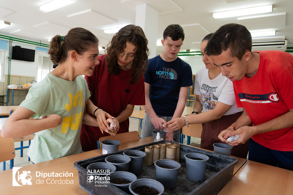 Cinco jóvenes participan en un taller de plantación en un aula, preparando macetas con tierra y semillas alrededor de una mesa. En la parte inferior aparecen los logotipos de la Diputación de Córdoba y de Albasur Plena inclusión Andalucía.